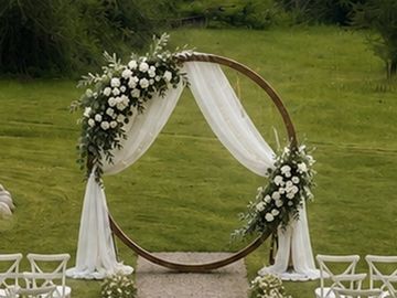 A circular wedding arch decorated with white flowers and sheer fabric on a grassy lawn.