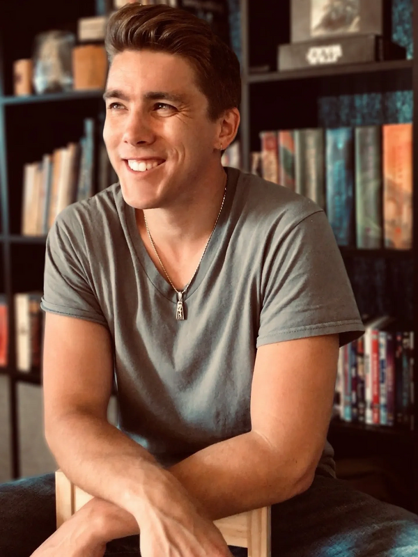 Dan Kelley sitting in a chair and smiling in front of a shelf filled with books, DVDs, & puzzles