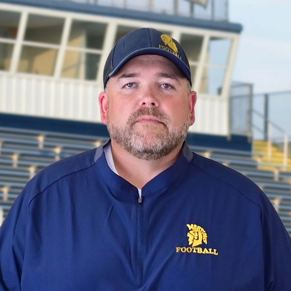 Man in a blue football jacket and cap with a serious expression.
