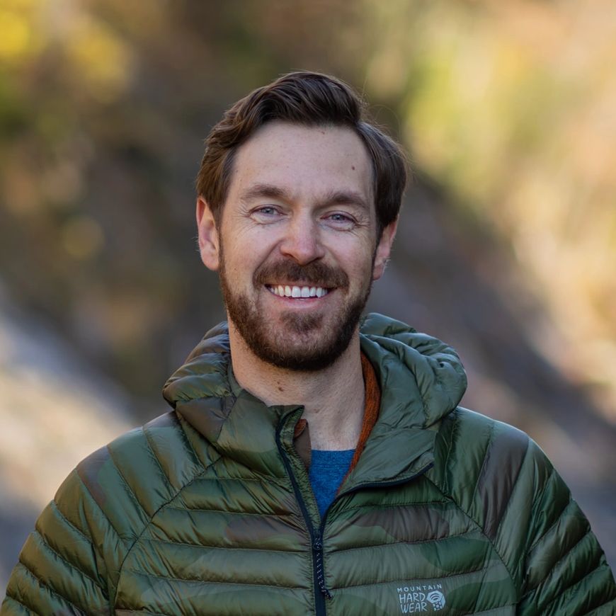 Smiling man in a green puffer jacket outdoors by a river.
