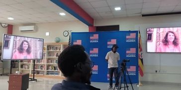 A man records a virtual interview in a room with Uganda and US flags.