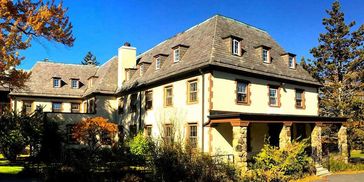 A large, beige mansion with a tiled roof surrounded by trees under a clear blue sky.