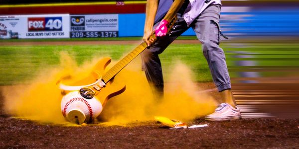 Person hitting a giant baseball with an electric guitar at a field.