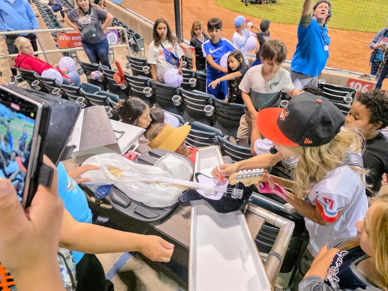 Woman in a baseball stadium signing a guitar for kids.