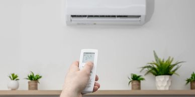 Hand holding remote aimed at an air conditioner on a white wall.