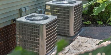 Two outdoor air conditioning units installed beside a house on a paved area.