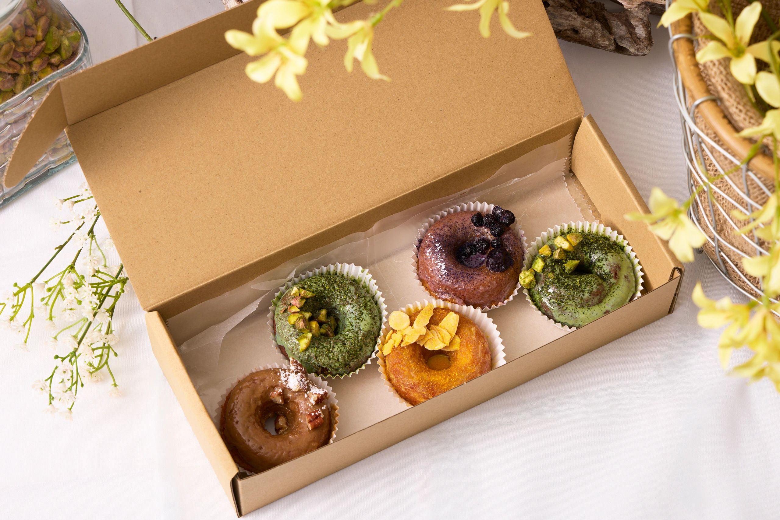 Assorted gourmet donuts in a cardboard box surrounded by flowers.