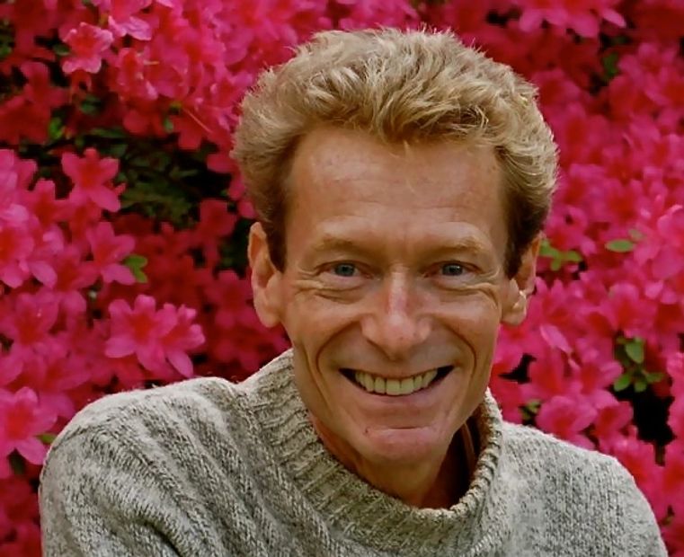 Smiling man with glasses in a gray sweater against vibrant red flowers.