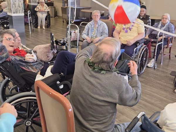 Seniors in wheelchairs play beach volleyball indoors with a colorful ball.