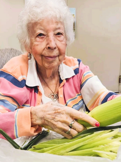 Elderly woman peeling fresh corn indoors, wearing a colorful striped sweater.