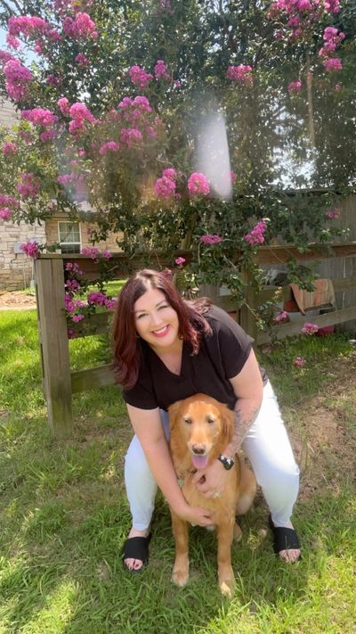 Woman smiling and hugging a golden retriever under blooming pink flowers.
