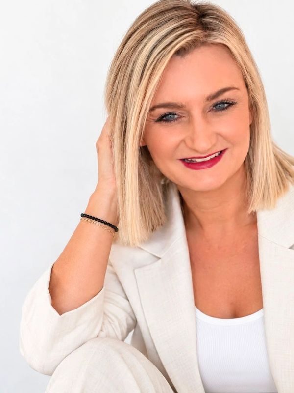 Smiling blonde woman in light suit touching her hair.