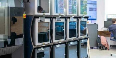 A row of 3D printers in a modern workspace with a person working in the background.
