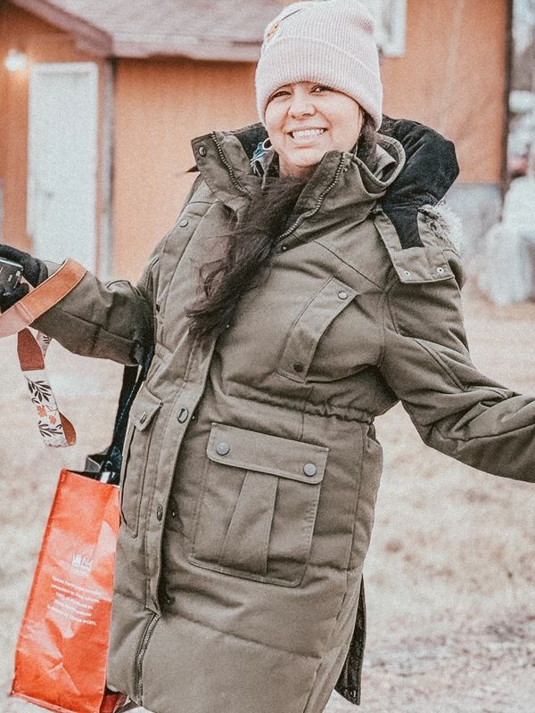 Robyn in winter coat joyfully posing outdoors with a camera and red bag.