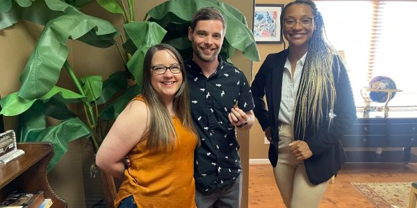 Three friends smiling indoors with plants in the background.