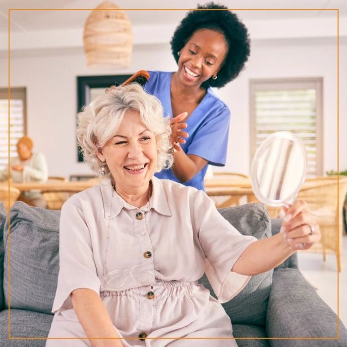 A caregiver happily combs an elderly woman's hair as she admires herself in a hand mirror.