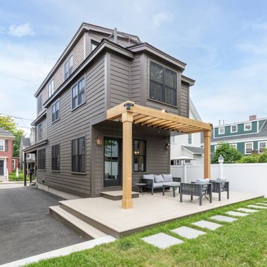 Modern gray house with a wooden pergola and outdoor seating on a sunny day.
