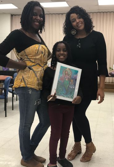 Three smiling females pose, with a young girl holding a framed mermaid artwork.