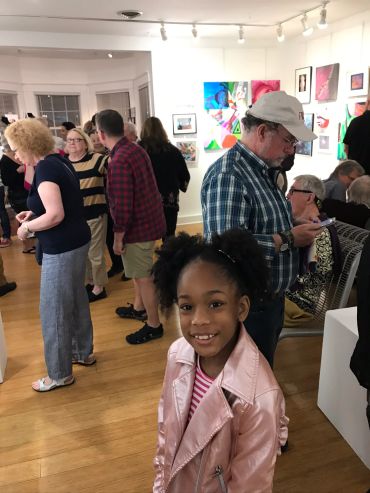 A smiling young girl in a pink jacket at an art gallery event.