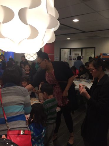 A lively indoor gathering with adults and children engaging in crafts under bright lighting.