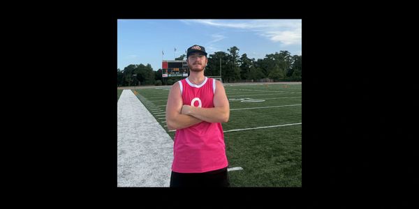 Man in a pink sleeveless shirt stands on a football field with arms crossed.