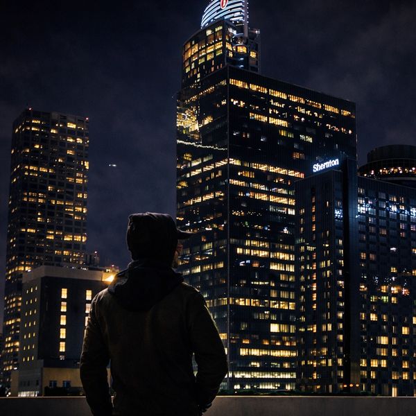 Person in hoodie gazing at illuminated skyscrapers in the night cityscape.