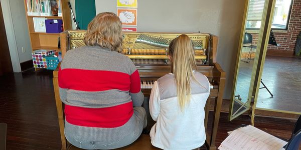 A student and piano teacher sit together on the piano bench. 