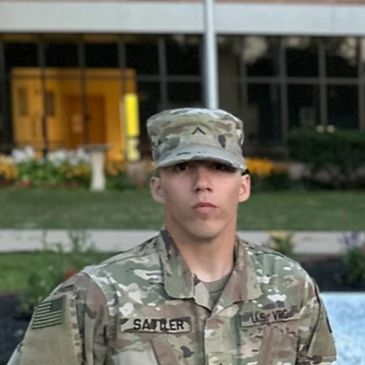 A soldier in camouflage uniform stands outdoors in front of a building.