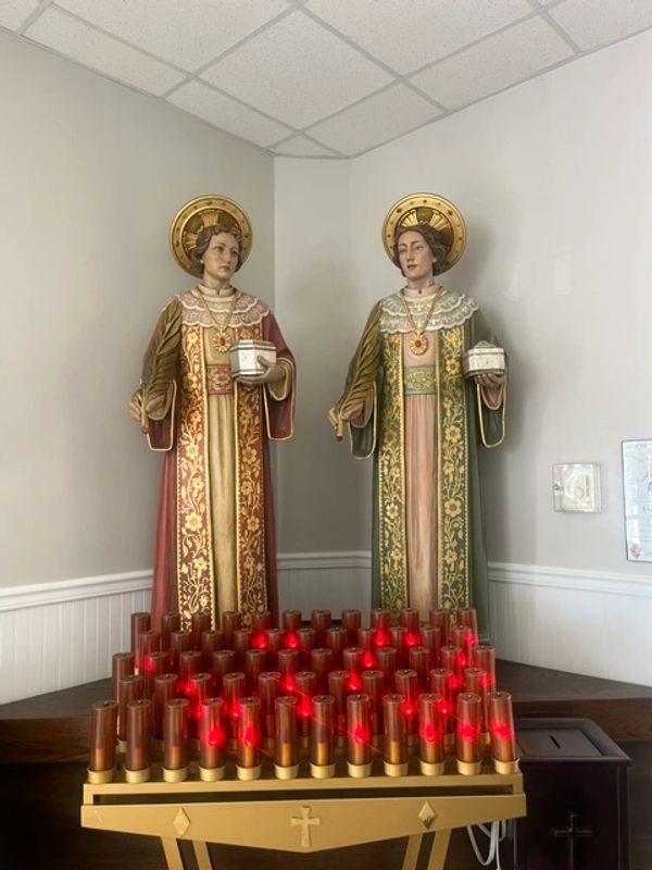 Two religious statues with halos stand behind lit red candles in a corner.