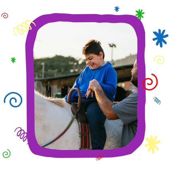 A joyful boy rides a white horse with adult support outdoors.