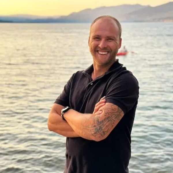 Smiling man with tattooed arm stands by a calm sea at sunset.