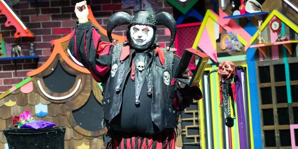 Performer in jester costume on stage with colorful houses.