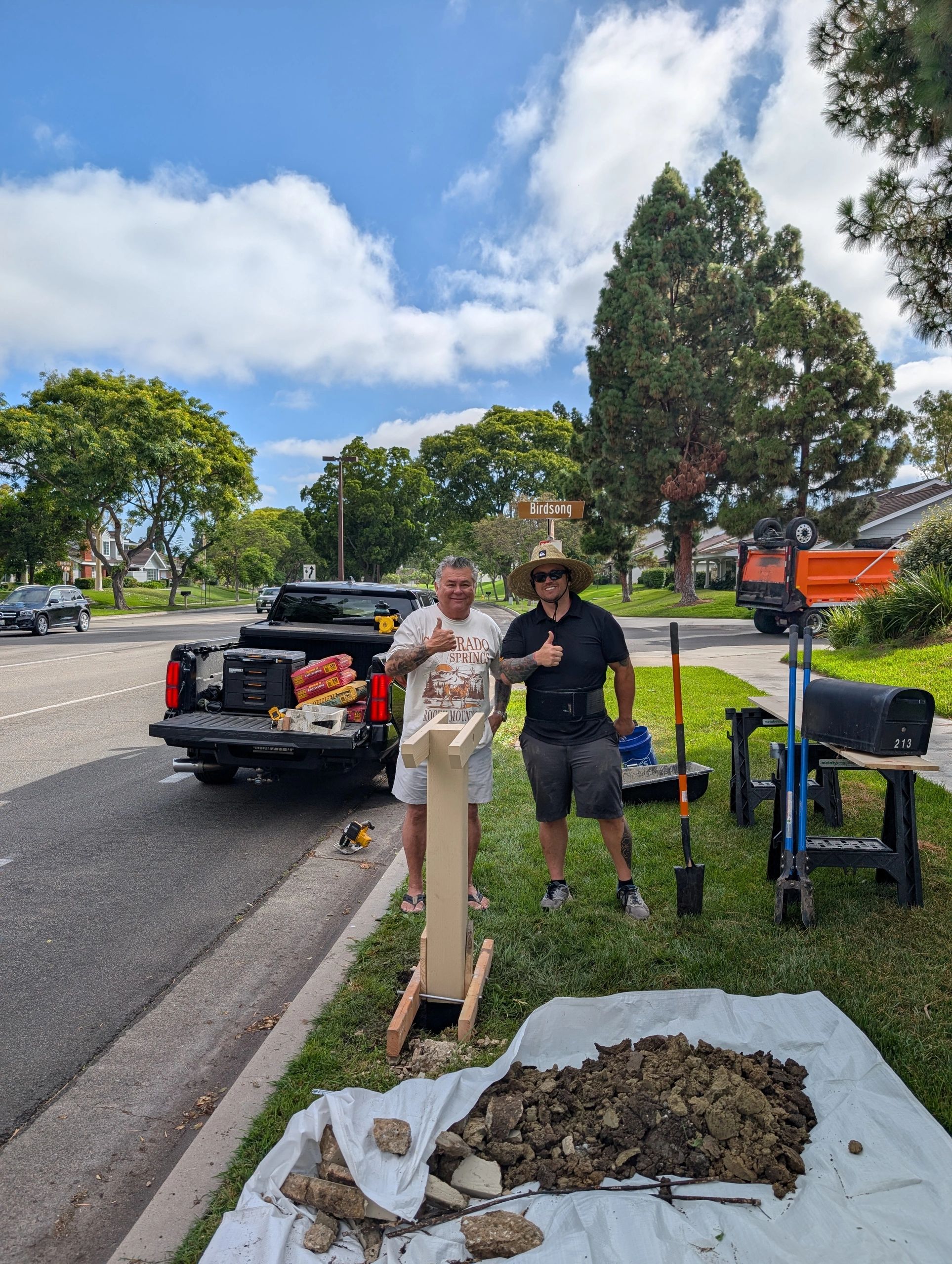 Two men giving thumbs up next to a newly installed wooden post on a sunny suburban street.