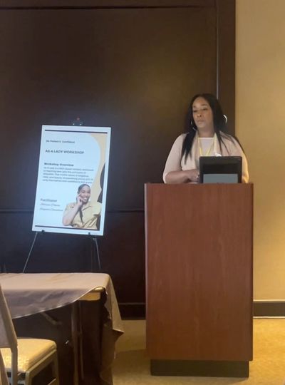 Woman presenting at a workshop behind a podium with an informational poster nearby.