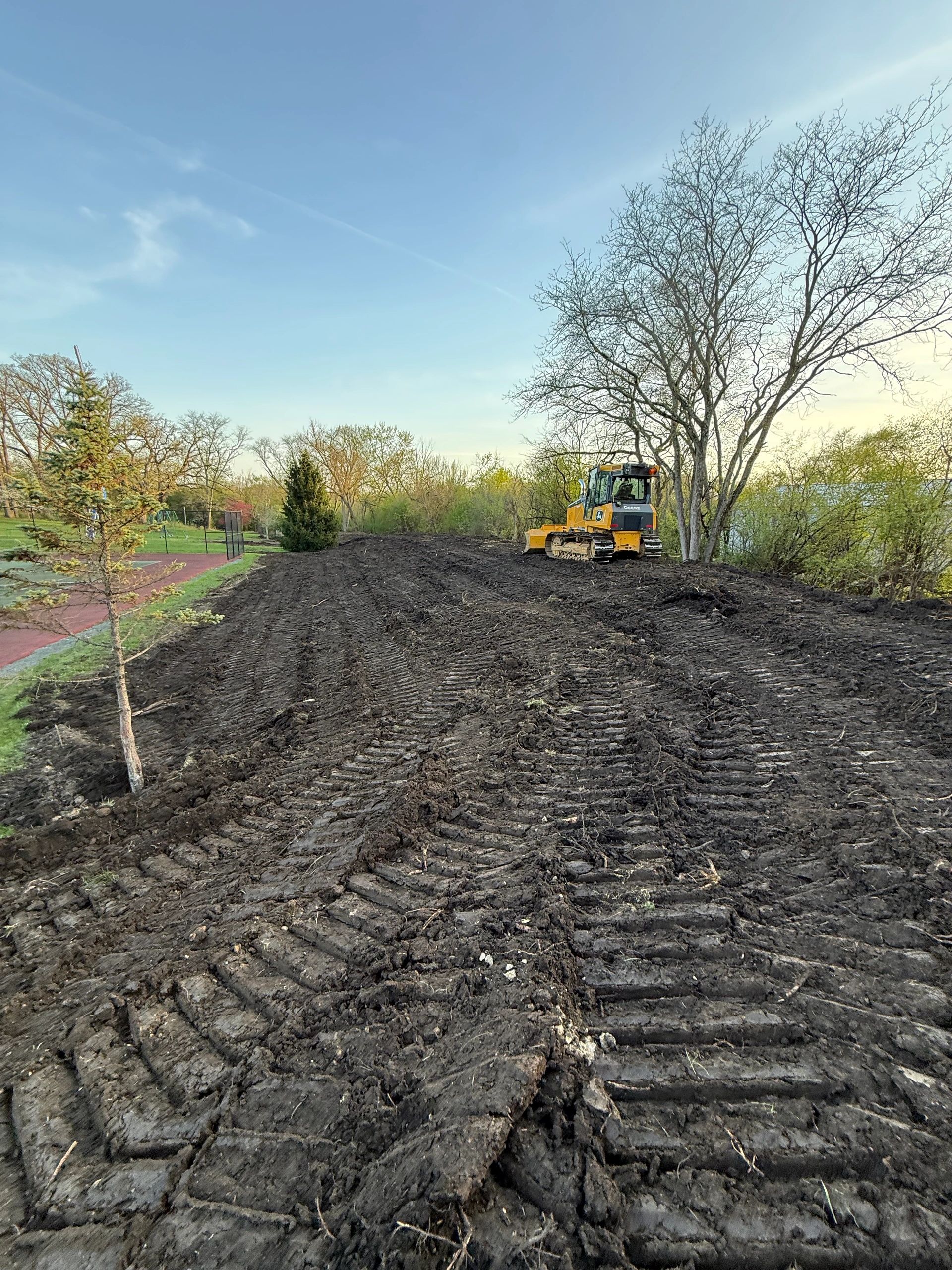 A yellow bulldozer on freshly plowed soil near trees and a tennis court.