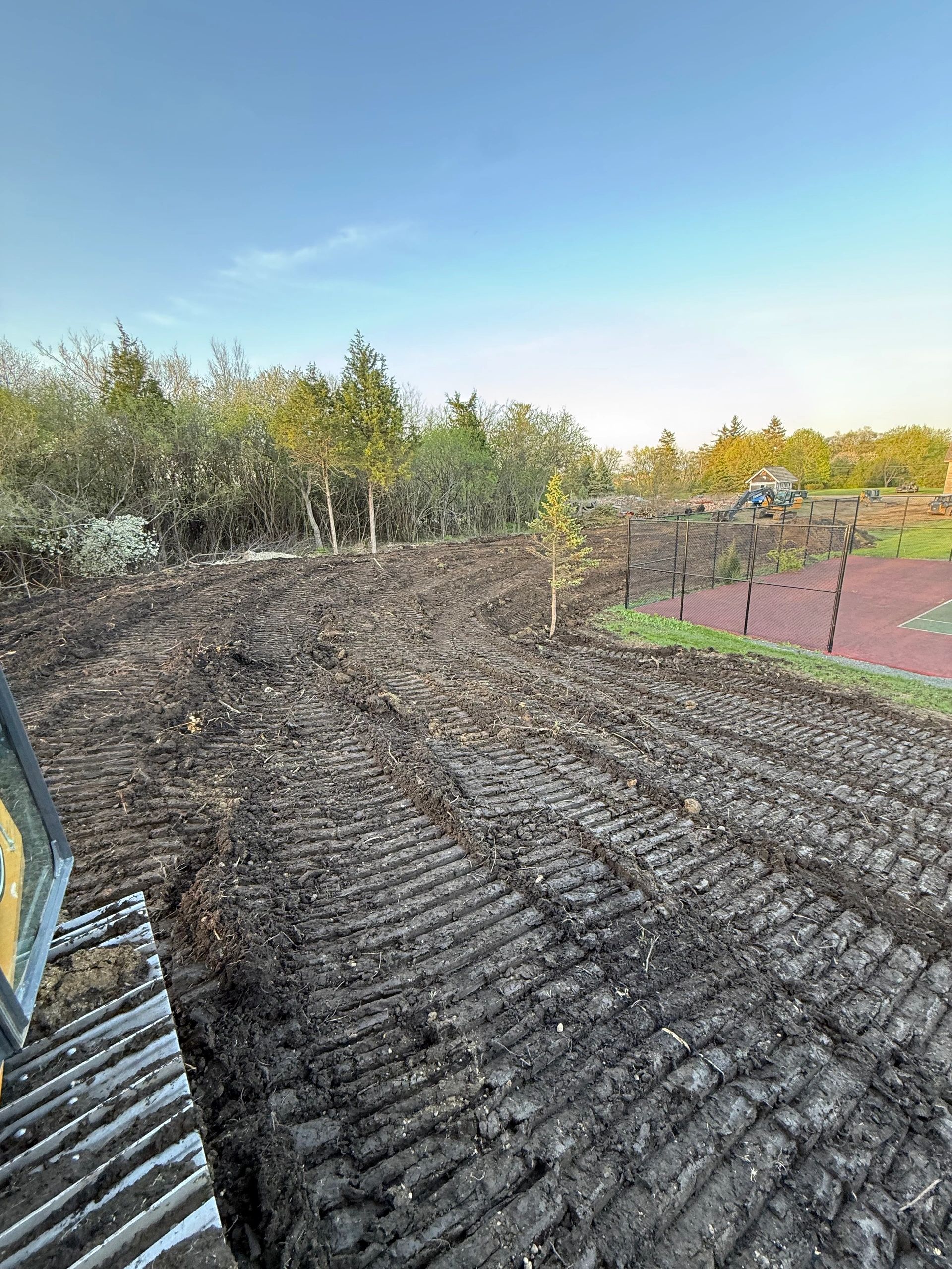 Freshly cleared land with tractor tracks near a fenced tennis court.