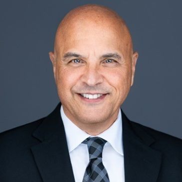 Smiling bald man in a suit and tie against a plain dark background.