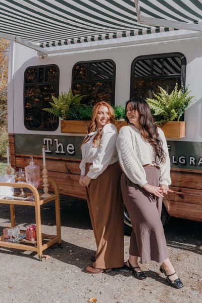 Two women smiling and posing in front of a plant-decorated trailer.