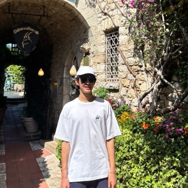 A person in a white cap and shirt stands outside a stone building with flowers.