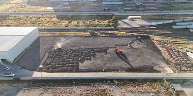 Aerial view of a large pile of dark material with a vehicle and sprinklers on an industrial site.