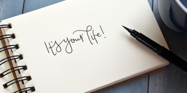 This is a notepad with "It's your life" written and sitting on a table with an ink pen placed on top.
