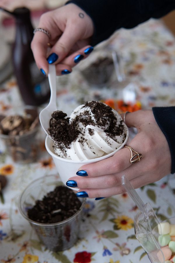 Hand with blue nails scooping cookie crumbs onto soft-serve ice cream.