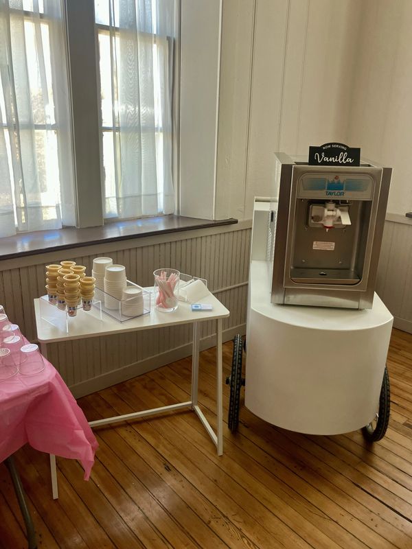 Vanilla soft serve machine with cones, cups, and spoons on a table.