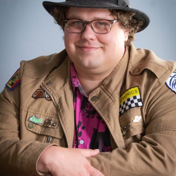 Man wearing glasses, hat, and a jacket with patches, smiling.