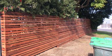 Modern wooden fence with greenery behind it on a sunny day.
