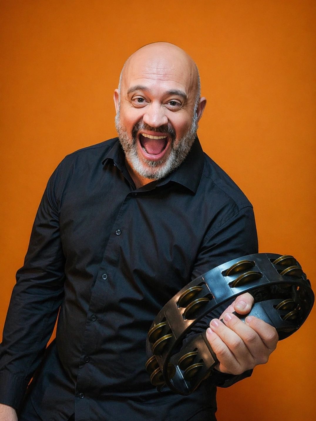 Man with a beard joyfully holding a tambourine against an orange background.
