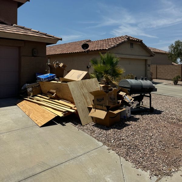 Pile of wooden boards, cardboard boxes, and a grill outside a house on a sunny day.