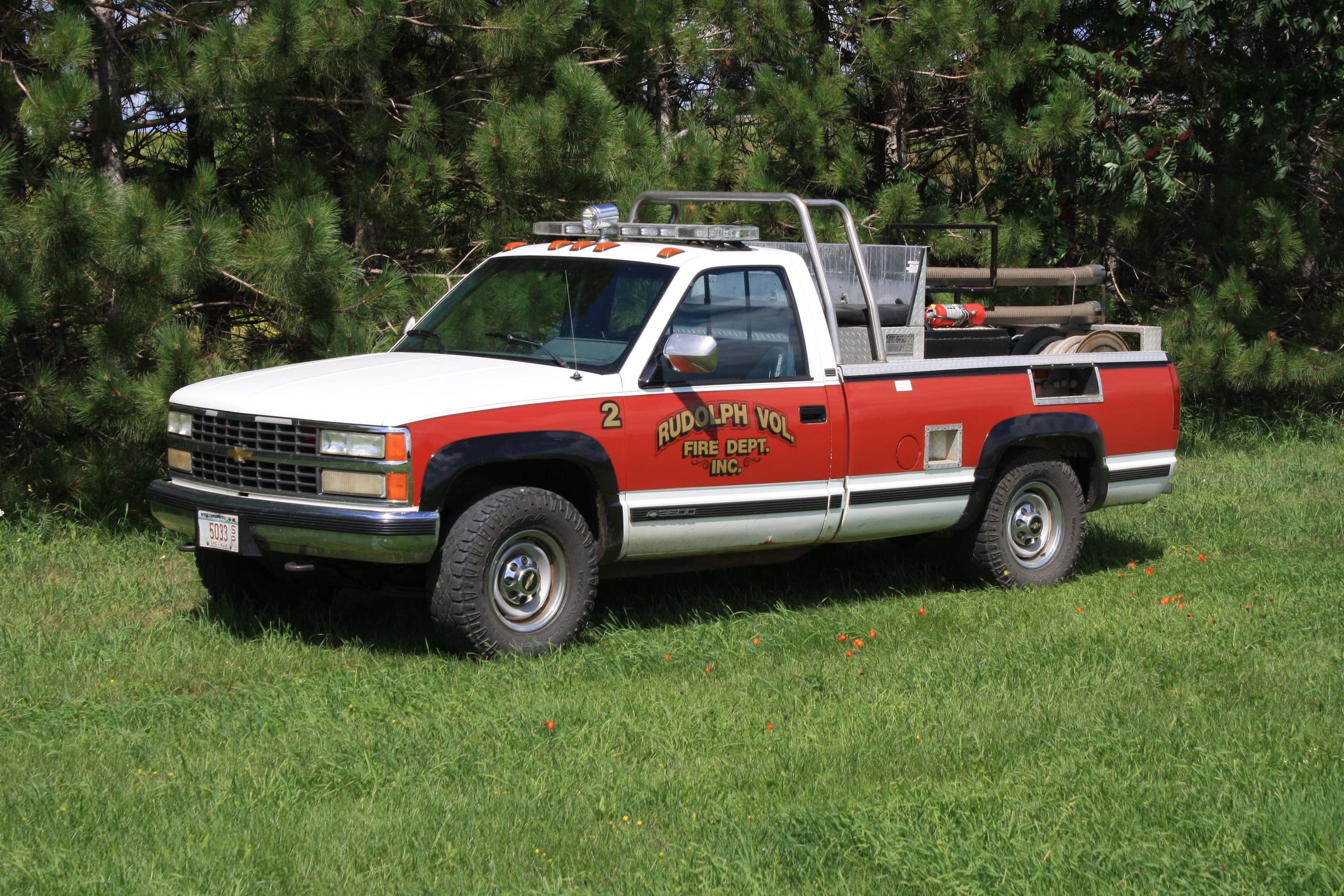 Red and white fire department pickup truck parked on grass near trees.