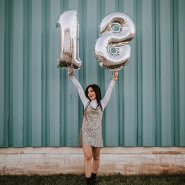 A black haired young girl holding up two baloons with a 18 on 