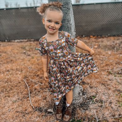 Smiling little girl in a floral dress and boots posing by a tree.